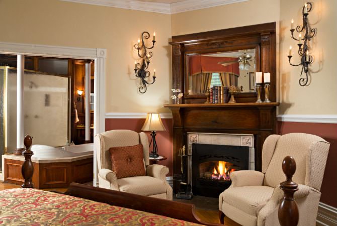 An elegant bedroom sitting area featuring two beige wingback chairs facing a glowing fireplace with a dark wood mantle. To the left, a large opening reveals a private bathroom with a wooden-paneled soaking tub.