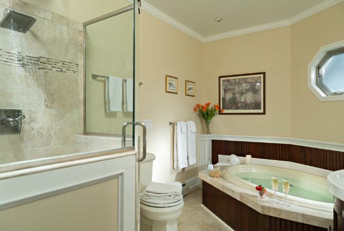 A brightly lit luxury bathroom featuring a large white spa tub with dark wood paneling, a glass-enclosed walk-in shower with a rainfall showerhead, and a small octagonal window.