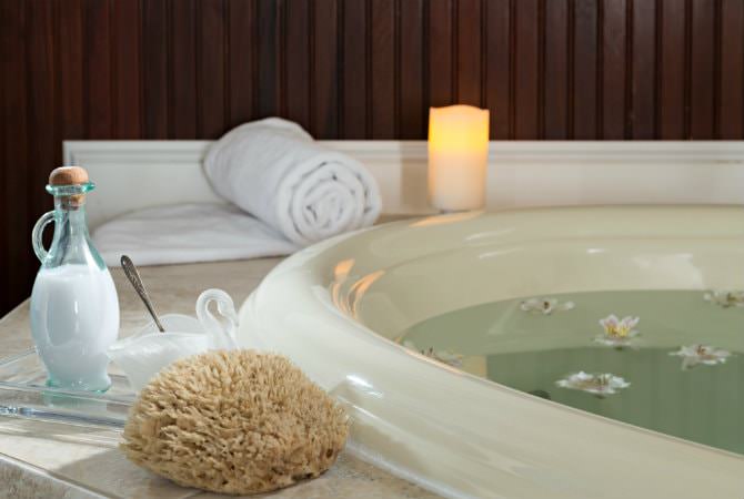 A close-up of a large white soaking tub filled with water and floating flower petals. On the marble edge, there is a natural bath sponge, a glass bottle of bath salts, a rolled white towel, and a glowing pillar candle against a dark wood-paneled wall.