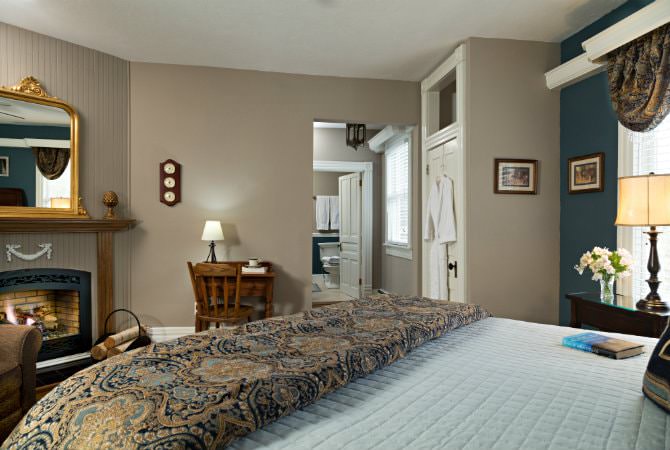 A cozy bedroom scene featuring a lit fireplace under a wooden mantle with a gold-framed mirror. In the foreground, the edge of a bed with a patterned blue and brown duvet is visible, and in the background, a small wooden desk sits near a doorway leading to a bright bathroom.