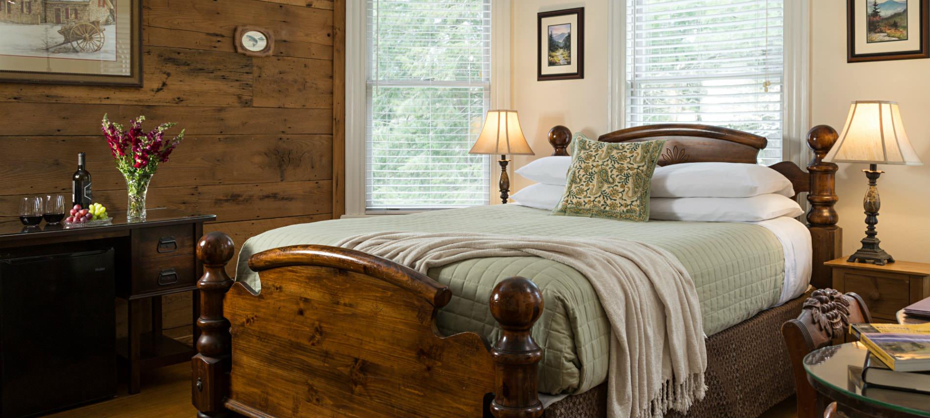 A cozy bedroom with a warm wood-paneled accent wall, featuring a high-backed wooden queen bed with green and white linens. Two bedside lamps provide warm light, and a vase of pink flowers sits on a dark wooden dresser in the foreground.