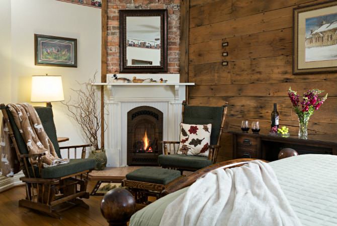 A cozy bedroom sitting area featuring two dark wooden rocking chairs with green cushions positioned in front of a white fireplace with a lit gas insert. The room has a rustic feel with a horizontal wood-plank accent wall, a brick chimney, and a wooden desk to the right adorned with a vase of red flowers and two glasses of wine.