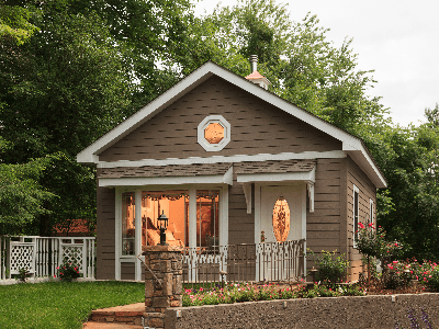 The Carriage House Suite's exterior at dusk, showing light glowing through a large bay window and an ornamental glass front door. The small, gray bungalow features white trim, a decorative round window above the entrance, and is surrounded by green trees and landscaped garden beds.