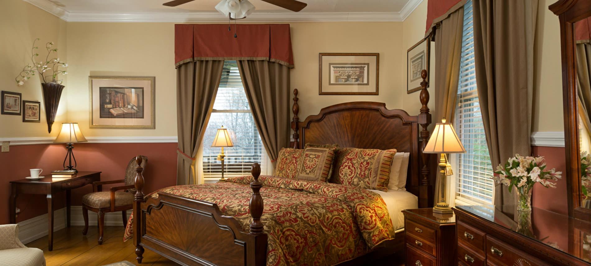 An elegant bedroom featuring a grand wooden four-poster bed with ornate gold and brown patterned linens, framed by large windows with heavy decorative drapery and a warm red accent wall.