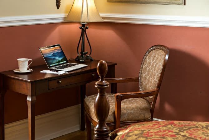 A classic dark wood writing desk with a matching upholstered chair tucked in the corner of a room with warm red and tan walls. On the desk sits a laptop showing a landscape image, a white coffee mug, and a lamp providing warm light.