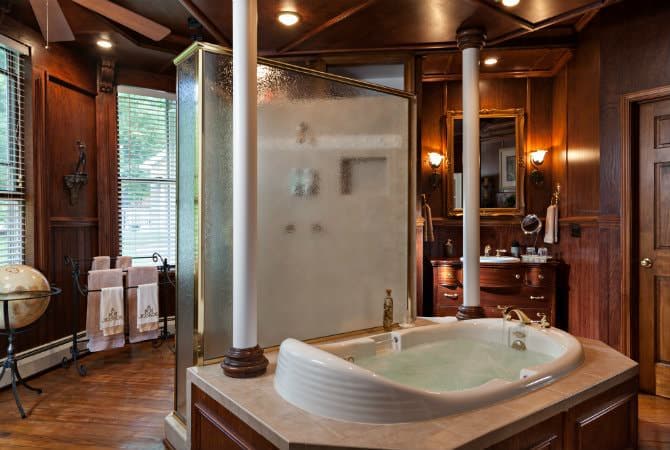 A luxurious wood-paneled bathroom featuring a large oval soaking tub in the foreground and a glass-enclosed walk-in shower. Two tall white columns flank the tub area, and a dark wood vanity with a large mirror is visible in the background.