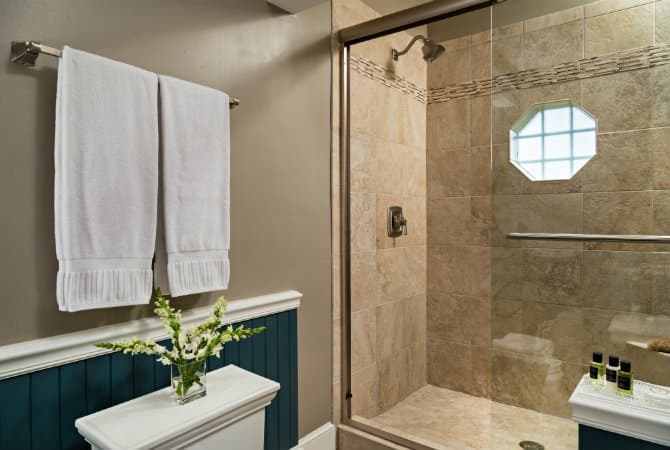 A bright, modern bathroom featuring a walk-in shower with beige tile and a small octagonal window. White towels hang on a silver rack against a gray wall with dark blue wainscoting, and a small vase of flowers sits on a white ledge in the foreground.