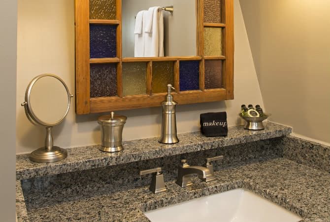 A close-up of a bathroom vanity area featuring a granite countertop and a white undermount sink. Above the counter, a wooden-framed mirror is decorated with squares of blue, yellow, and orange stained glass, and a small glass shelf holds silver canisters, a soap dispenser, and a small black clock.