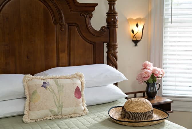 A close-up of a high-backed wooden bed with white pillows and a decorative floral throw pillow. In the foreground, a straw hat rests on the bed, while a small side table holds a vase of pink roses next to a window with white blinds.