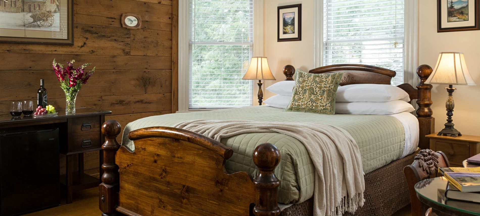 A cozy bedroom with a warm wood-paneled accent wall, featuring a high-backed wooden queen bed with green and white linens. Two bedside lamps provide warm light, and a vase of pink flowers sits on a dark wooden dresser in the foreground.