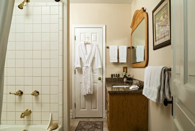 A classic bathroom featuring a white-tiled bathtub and shower combination with brass fixtures on the left. A white door with a hanging plush bathrobe stands in the center, next to a wooden vanity with a granite countertop, a large mirror, and neatly folded white towels.