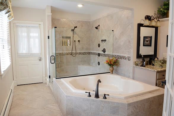 A luxurious white spa tub sits in the foreground of a bright bathroom, featuring a large glass-enclosed walk-in shower with multiple body jets and a rainfall showerhead in the background.