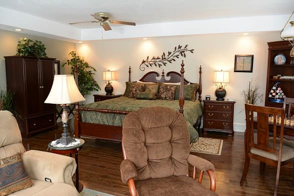 A classic, romantic suite featuring a dark wood four-poster king bed with green patterned bedding, two comfortable upholstered armchairs, and a matching wooden armoire and desk.