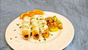 Three taquitos drizzled with sour cream, served with a colorful vegetable salad and orange slices on a decorative plate.