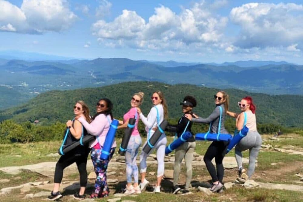 A group of seven women poses together on a scenic mountain overlook, each holding a yoga mat, with rolling hills in the background under a blue sky.