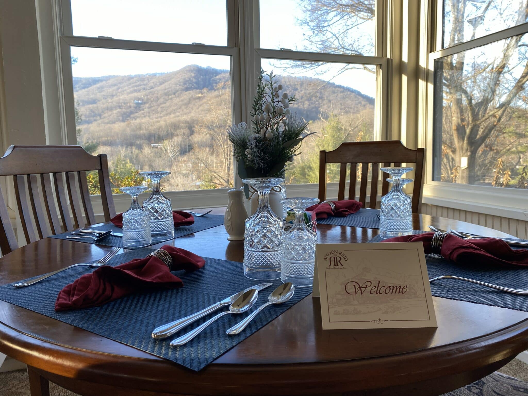 A beautifully set dining table with crystal glassware and red napkins, featuring a "Welcome" sign and a scenic view of mountains outside. A beautifully set dining table with crystal glassware and red napkins, featuring a "Welcome" sign and a scenic view of mountains outside.