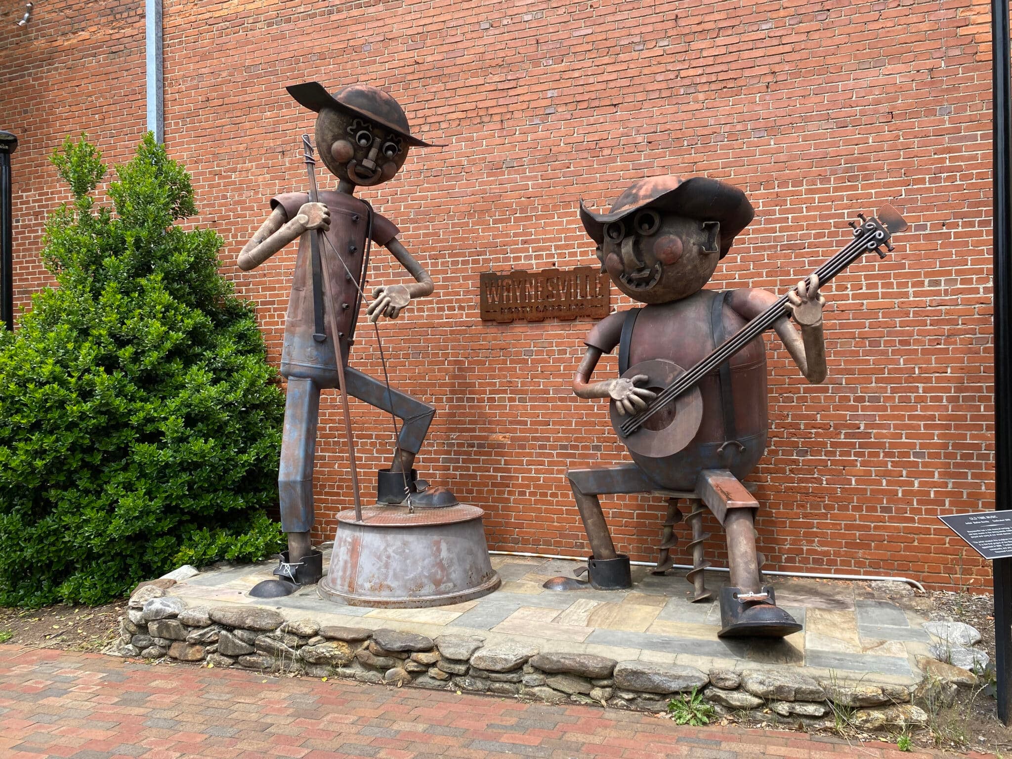 Two large metal sculptures of musicians playing a fiddle and banjo in front of a brick wall. Two large metal sculptures of musicians playing a fiddle and banjo in front of a brick wall.