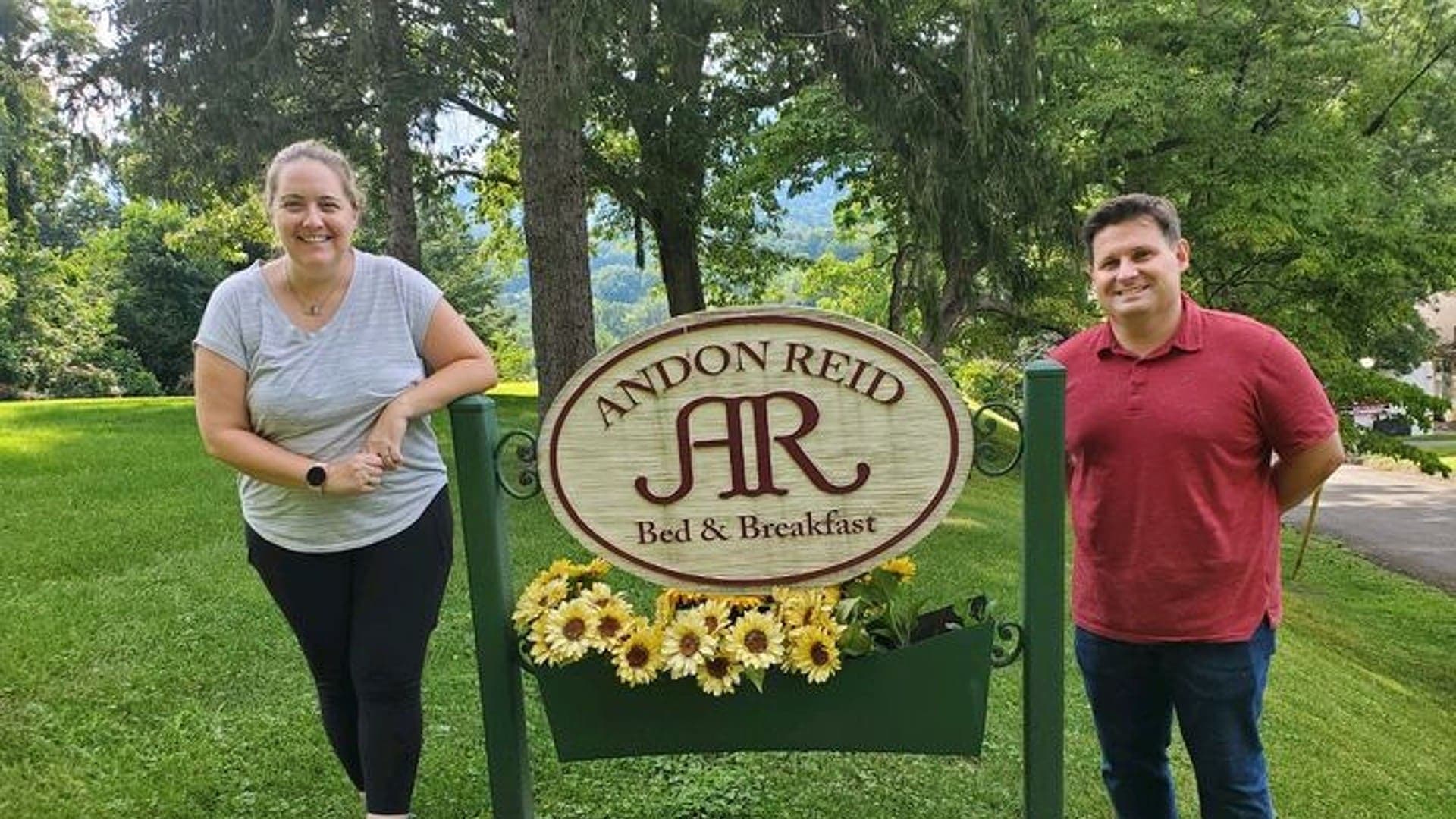 Two people stand beside a sign for Andon Reid Bed & Breakfast, surrounded by greenery and flowers. Two people stand beside a sign for Andon Reid Bed & Breakfast, surrounded by greenery and flowers.