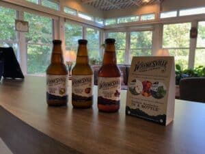 Three bottles of craft beverages and a box of assorted snacks on a wooden table in a sunny, plant-filled room.