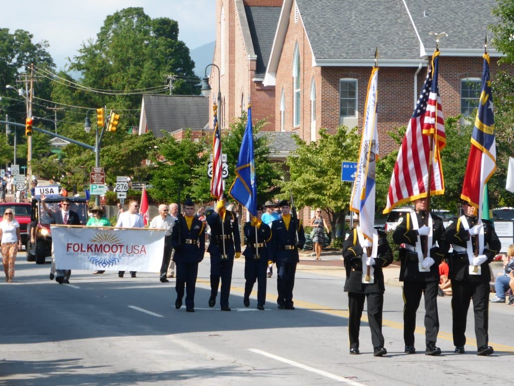 A parade featuring color guard members and participants holding a banner for "FolkMoot USA" on a sunny day.