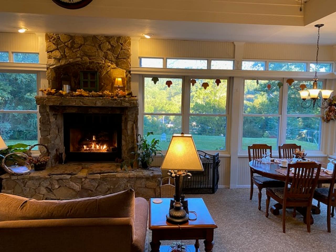 Cozy living room with a stone fireplace, a dining table, and large windows overlooking greenery.