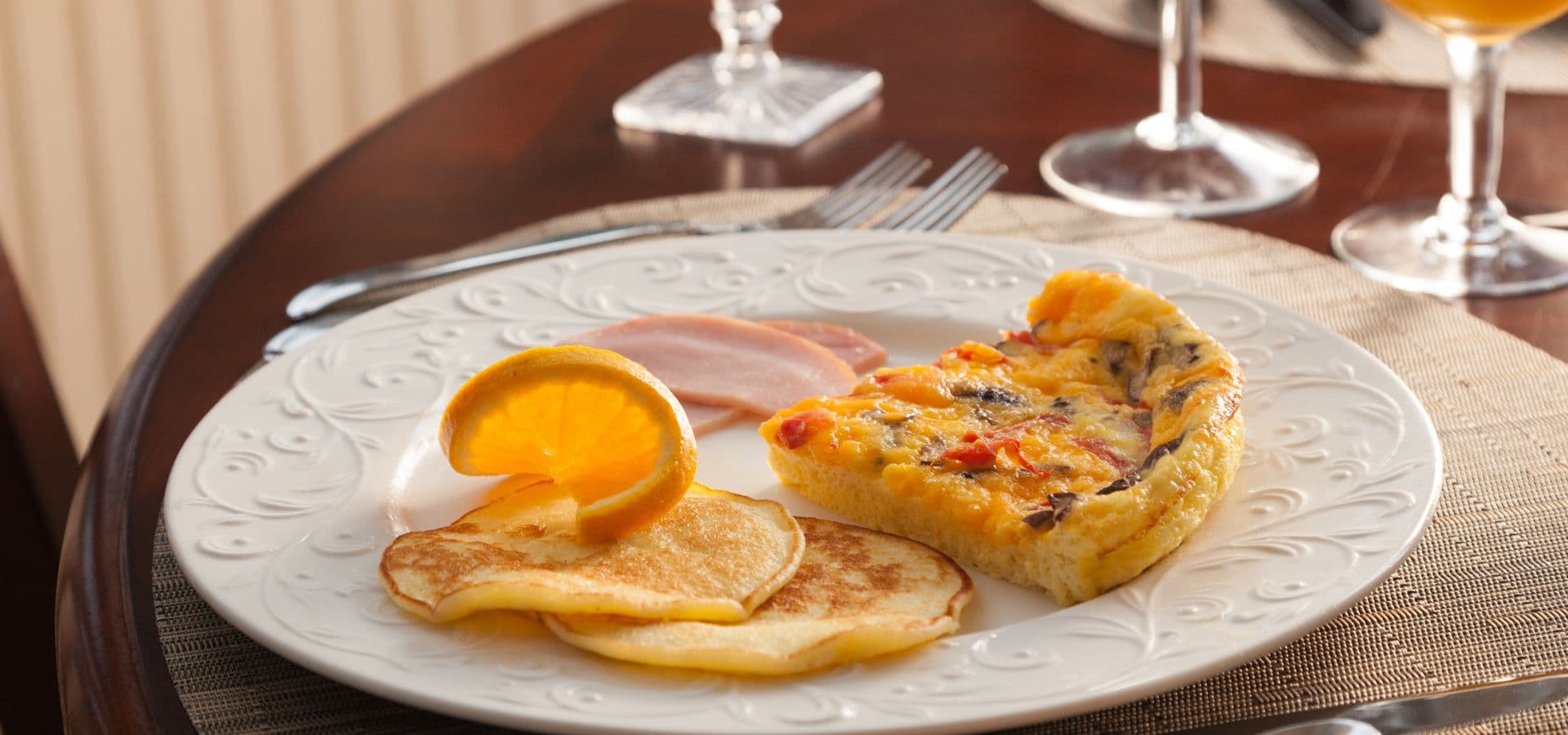 A plate featuring pancakes, a frittata, a slice of ham, and an orange slice on a dining table.