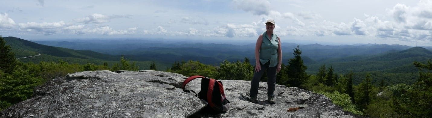 A person stands on a rocky outcrop overlooking a lush, mountainous landscape under a partly cloudy sky.