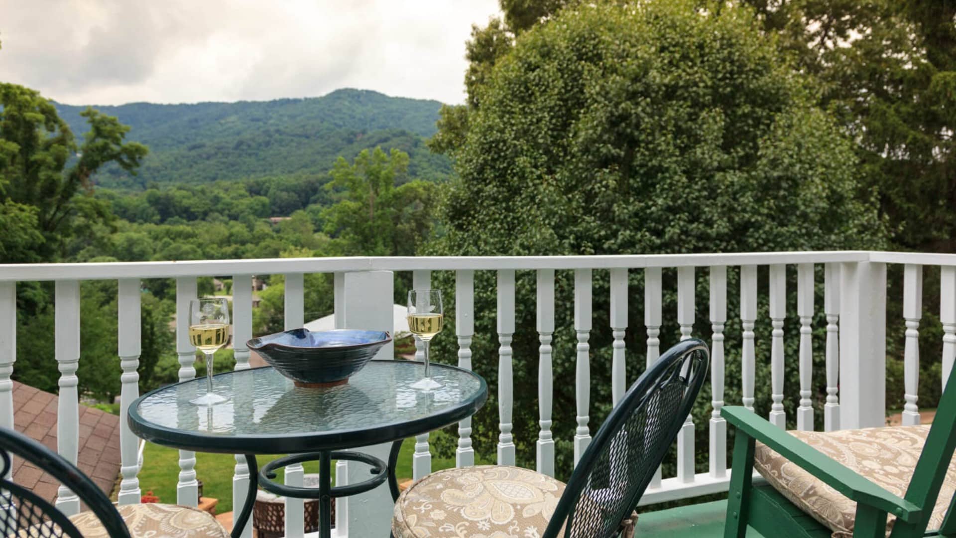 A balcony with a glass table and two glasses of white wine overlooks green hills and trees.