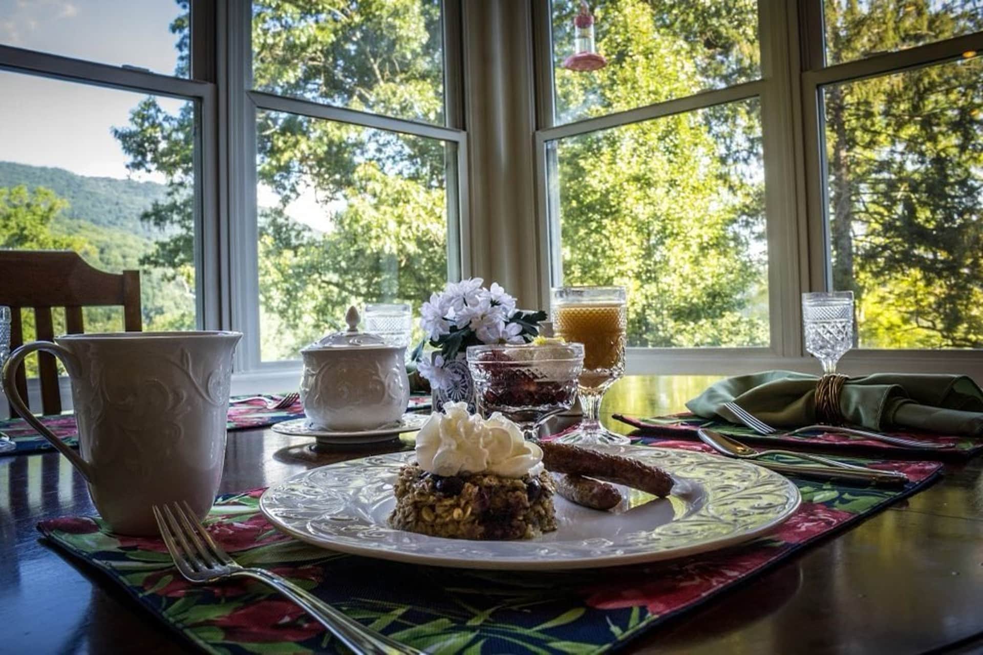 A beautifully set dining table with breakfast items, including a dessert, coffee, and juice, against a backdrop of large windows showcasing green trees.