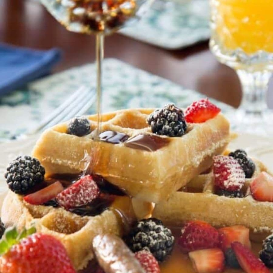 Stacked waffles topped with fresh berries and syrup, with a glass of orange juice in the background.