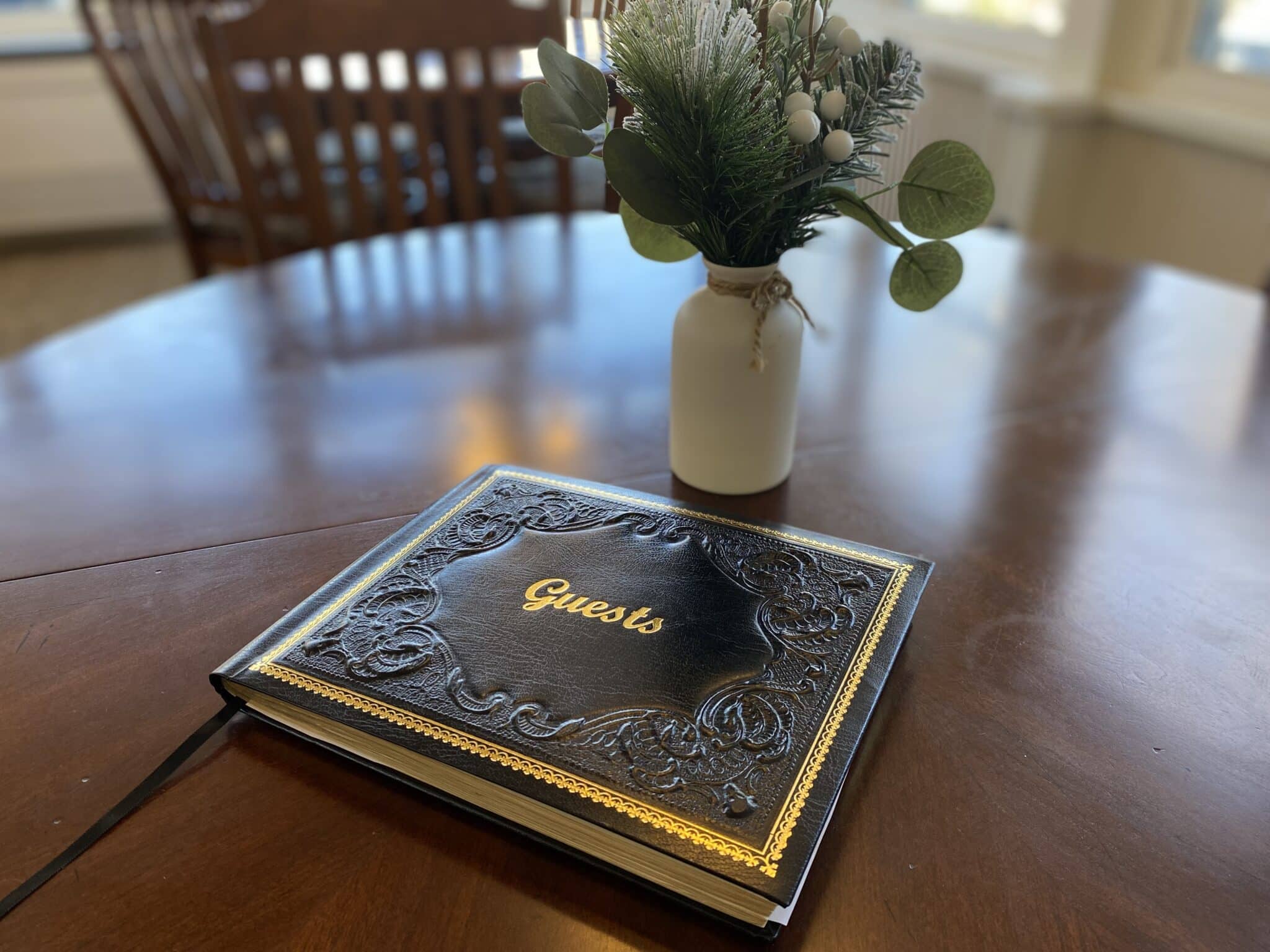 A decorative guest book sits on a wooden table next to a small vase with greenery. A decorative guest book sits on a wooden table next to a small vase with greenery.