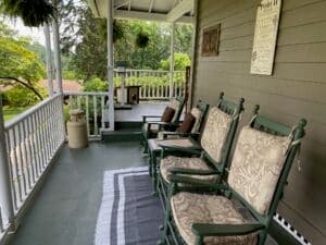A front porch featuring rocking chairs, a rug, and greenery in the background.