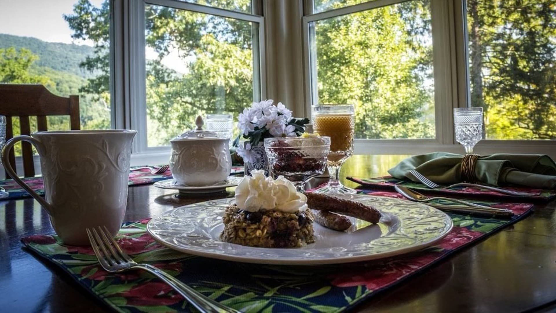 A beautifully set dining table with a plate of dessert, coffee, and floral arrangements, overlooking a scenic view.