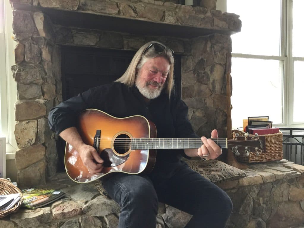 A man with long hair and a beard plays an acoustic guitar by a stone fireplace.