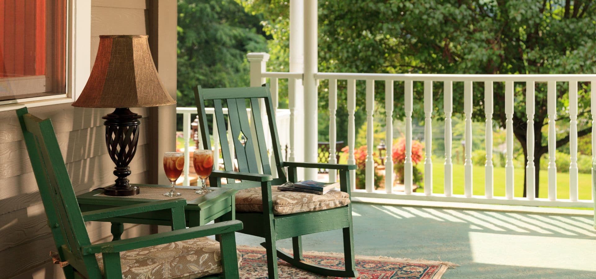 A cozy porch featuring two green rocking chairs, a lamp, and two drinks on a side table, surrounded by greenery.