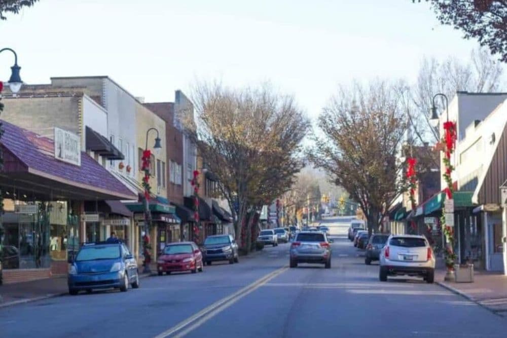 Main street lined with shops and decorated for the holidays, featuring parked cars and bare trees.