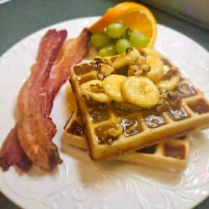 A plate of waffles topped with bananas and walnuts, accompanied by crispy bacon, green grapes, and a slice of orange.