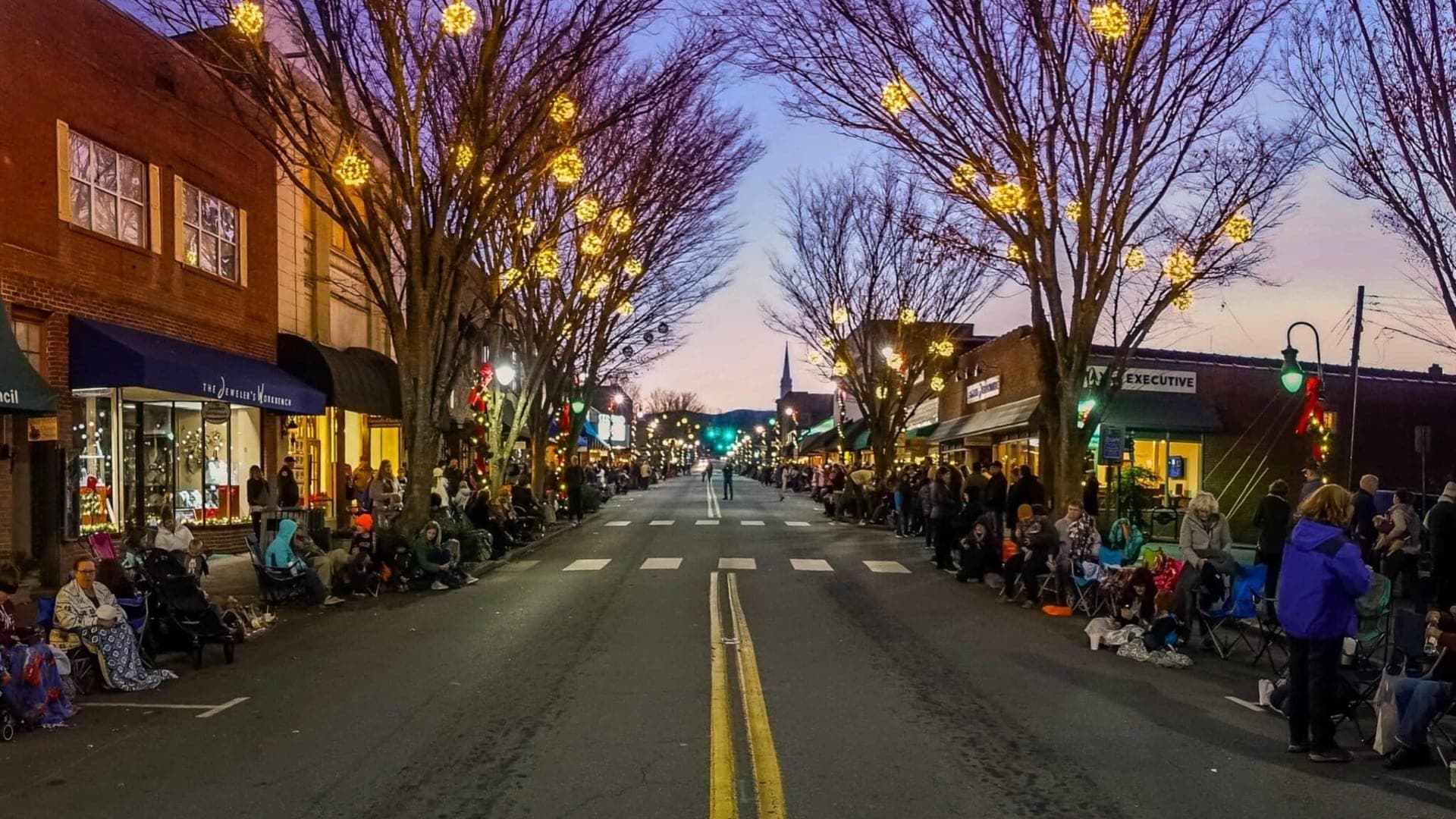 A festive parade street lined with spectators and decorated trees at dusk.