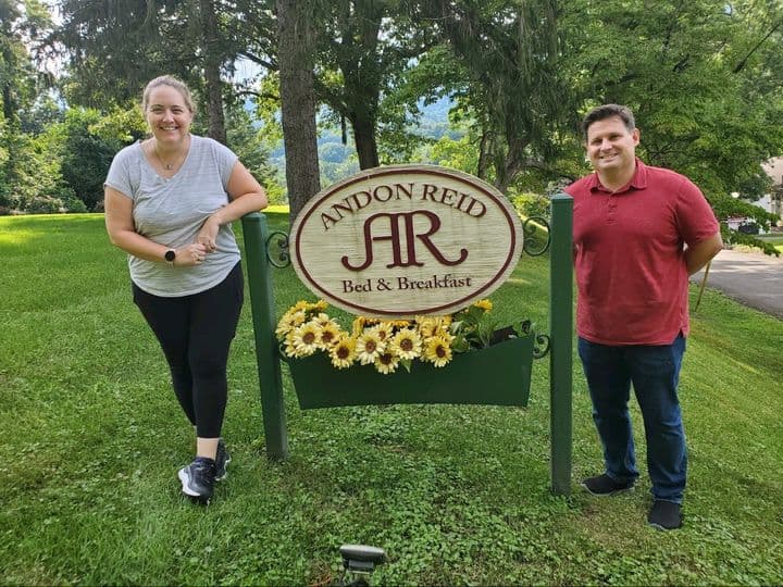 Two people stand beside a sign for Andon Reid Bed & Breakfast, surrounded by greenery and sunflowers.