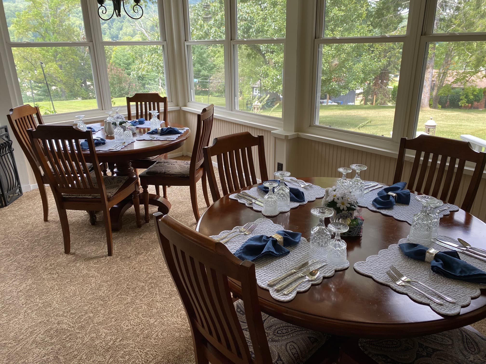 A sunlit dining room with two elegantly set tables featuring blue napkins and floral centerpieces.