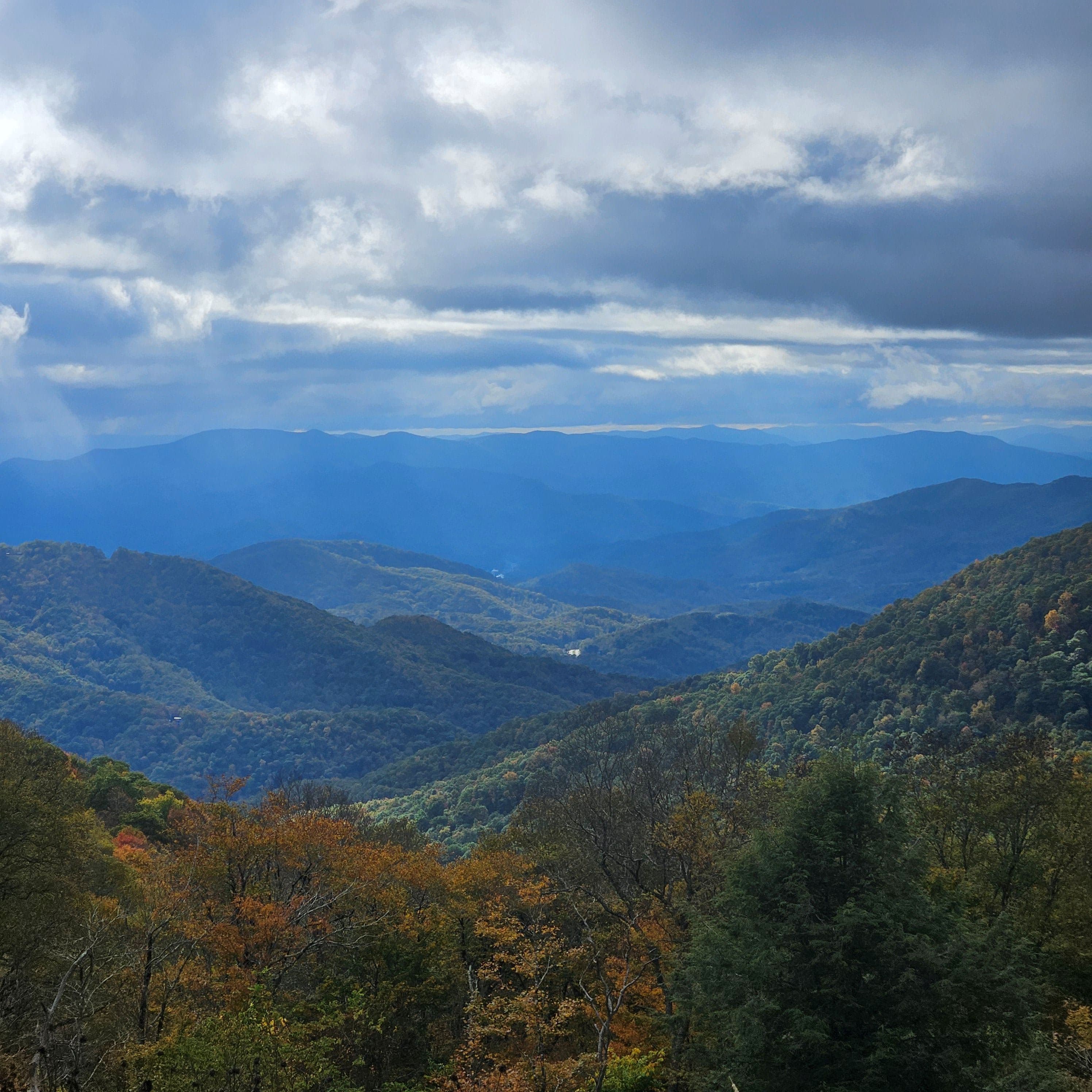 A panoramic view of layered mountains under a cloudy sky with hints of autumn foliage.
