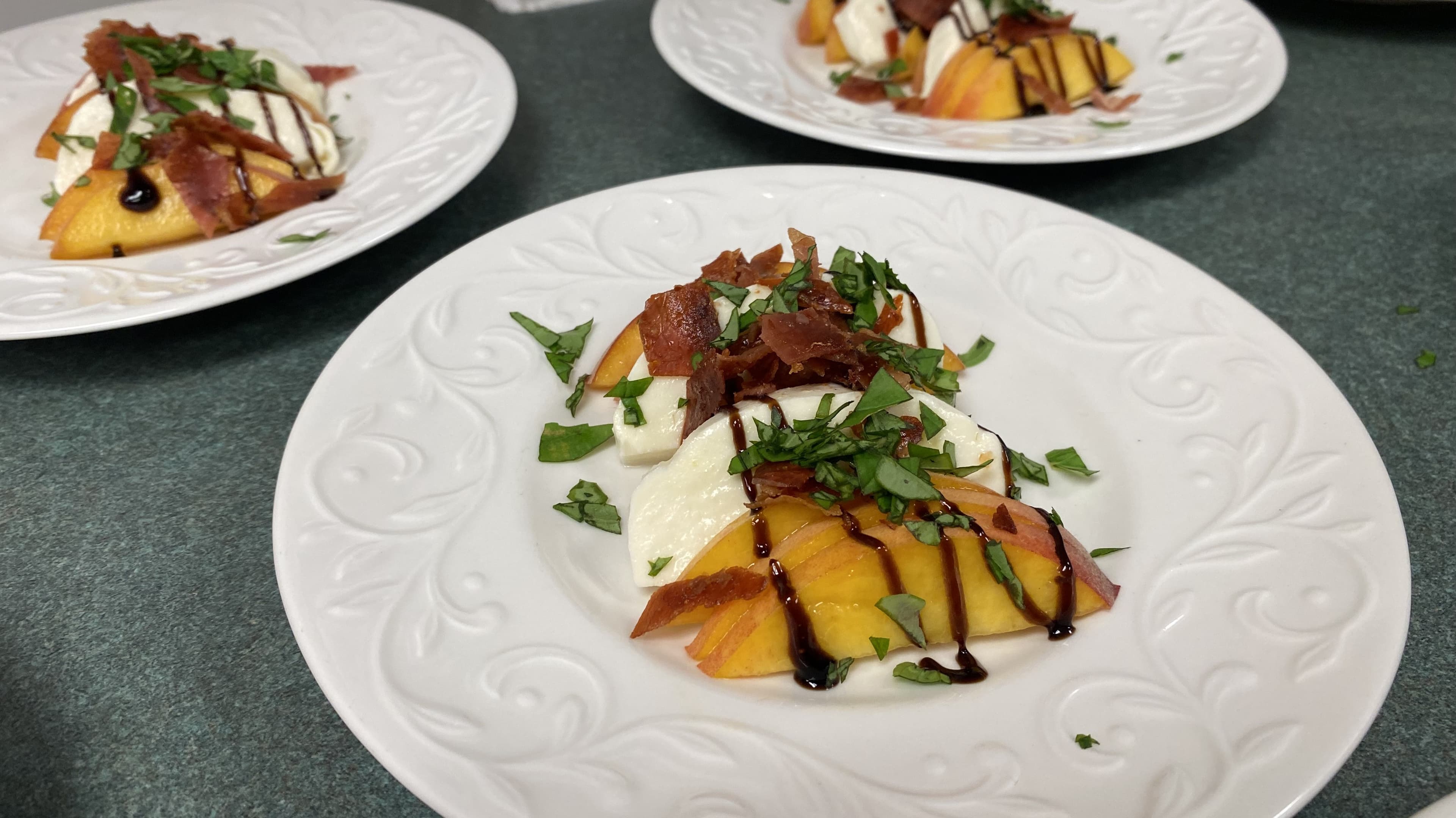 A beautifully arranged dish featuring sliced peaches, creamy cheese, crispy bacon, and fresh basil drizzled with balsamic glaze on a decorative plate.