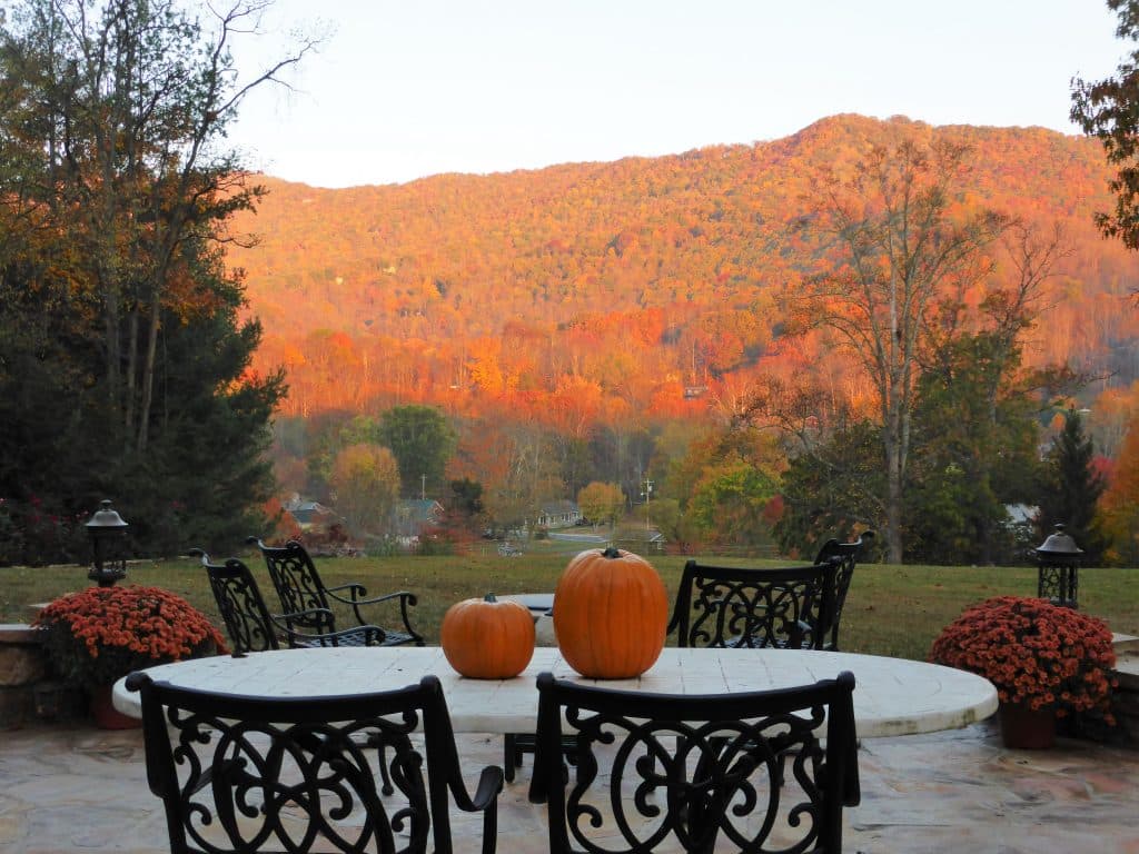 A scenic view of autumn hills with vibrant foliage, featuring a stone table adorned with two pumpkins and surrounding chairs.