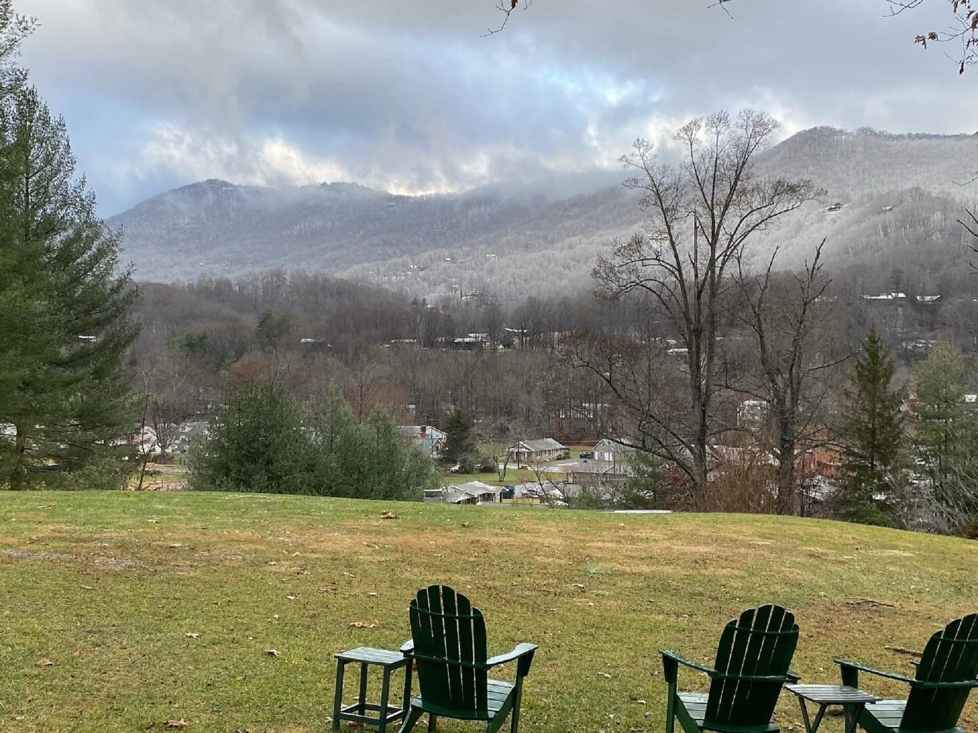 A serene view of mountains and a small town from a grassy area with three green chairs in the foreground. A serene view of mountains and a small town from a grassy area with three green chairs in the foreground.