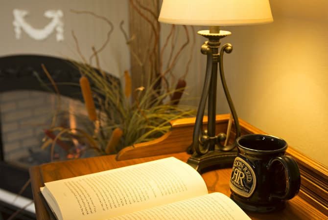 A close-up of an open book on a wooden desk, illuminated by a warm table lamp. Next to the book is a dark, branded ceramic mug, with a glowing fireplace and a potted plant softly blurred in the background.