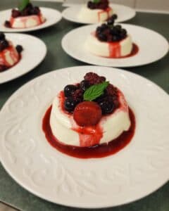 A creamy panna cotta topped with mixed berries and a raspberry sauce, served on a decorative white plate.