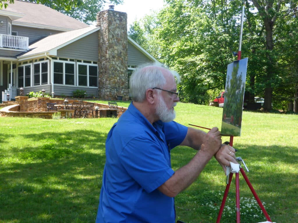 An elderly man with a beard paints on an easel in a lush, green backyard.