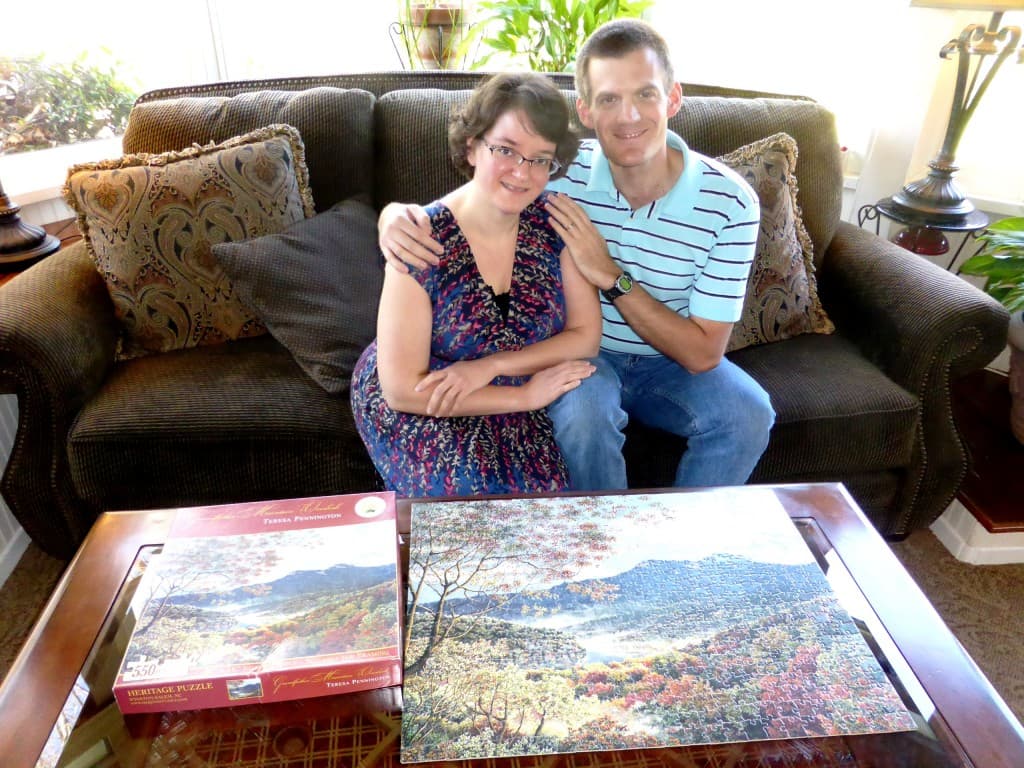 A couple sits together on a couch, smiling and posing beside a completed scenic puzzle and its original box.