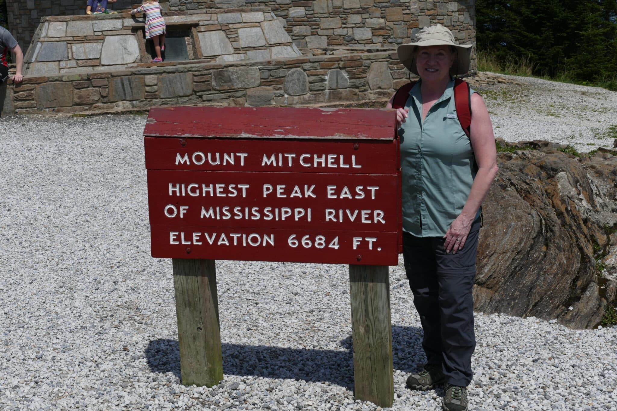 A woman stands next to a sign that reads "Mount Mitchell, Highest Peak East of Mississippi River, Elevation 6684 ft."