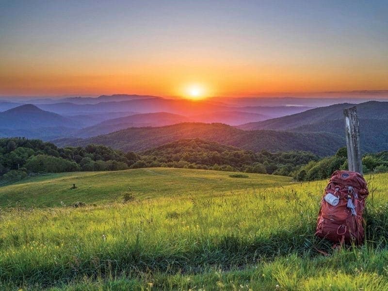 A backpack sits on a grassy hillside as the sun sets over rolling mountains.
